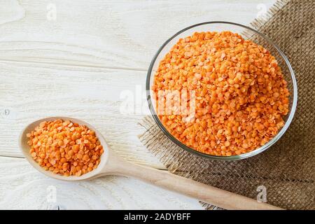 Getrocknete rote Linsen in einer Glasschale und Holzlöffel Sacktuch Tabelle Serviette über Weiß rustikal Tisch. Zutaten für vegetarische Suppe pürieren. Stockfoto