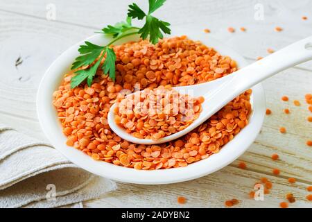 Nahaufnahme der getrocknete rote Linsen in einer weißen Schüssel mit Porzellan Löffel auf Holz- rustikalen Tisch. Zutaten für vegetarische Suppe pürieren. Das gesunde Essen. Stockfoto