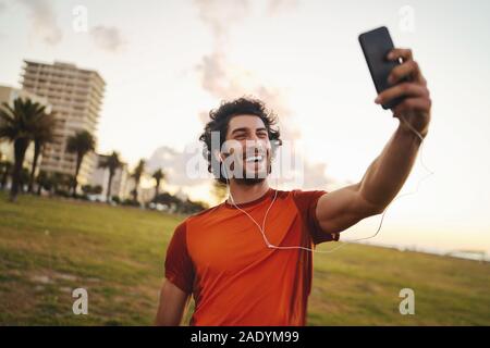 Lächelnd Portrait von einem kaukasischen passen junge Mann mit Kopfhörern in den Ohren die Videokonferenz von smart phone nach dem Training im Park - junge männliche Stockfoto