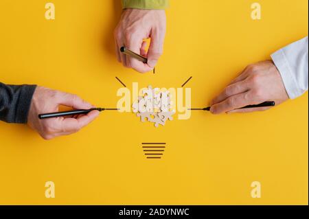 Drei männliche Hände zeichnen eine Glühbirne mit puzzle Stücke in der Mitte in einem konzeptionellen Bild von Vision und Teamarbeit. Auf gelben Hintergrund. Stockfoto