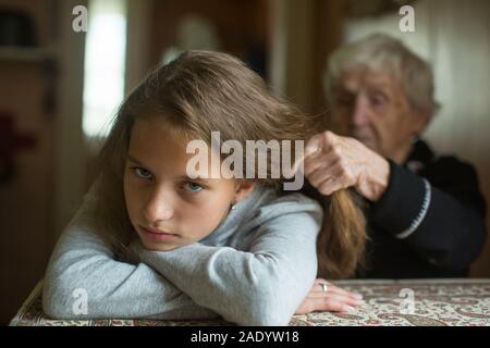 Verärgerte jugendlich Mädchen Winces an der Tatsache, dass ihre Großmutter Zöpfe Haar. Stockfoto