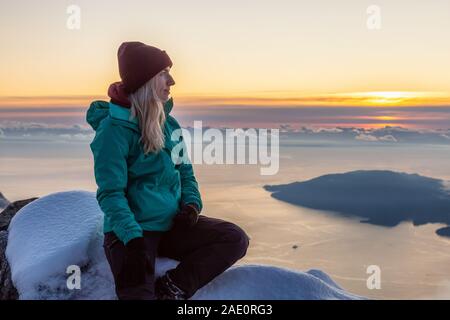 Abenteuerliche Blond kaukasische Mädchen sitzen auf einem Snow Mountain bei einem bunten Winter Sonnenuntergang abgedeckt. Auf St Mark's Summit, nördlich von Vancou genommen Stockfoto