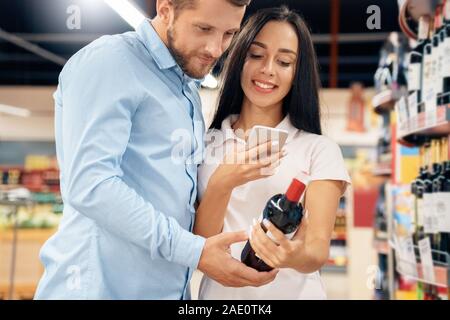 Die täglichen Einkäufe. Paar im Supermarkt zusammen an Alkohol Abteilung Auswahl Wein scannen QR-Code auf dem Smartphone Suchen Info freudige close-up Stockfoto