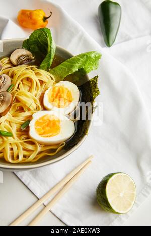 Traditionelle asiatische Ramen in der Schüssel in der Nähe von Stäbchen, Serviette und Zutaten auf graue Oberfläche Stockfoto