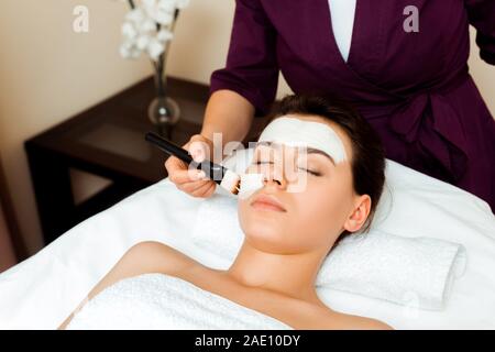 7/8-Ansicht der kosmetologin Anwendung Gesichtsmaske auf attraktive Frau in Spa Stockfoto