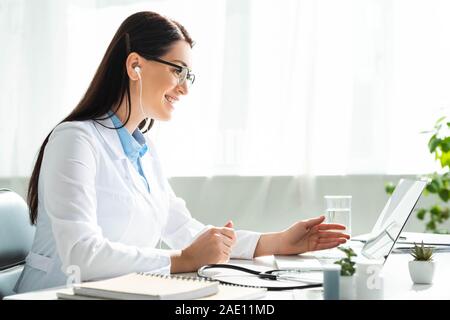 Positive Arzt der Ohrhörer in On-line-Konsultation mit Patienten auf Laptop in Klinik Büro Stockfoto