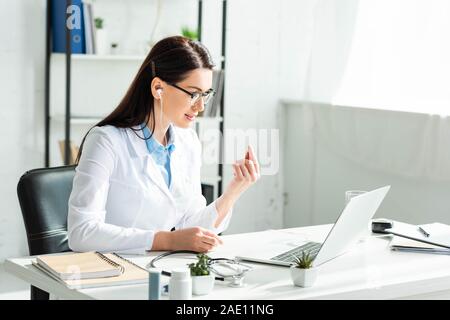 Weibliche positive Arzt der Ohrhörer in On-line-Konsultation mit Patienten auf Laptop in Klinik Büro Stockfoto