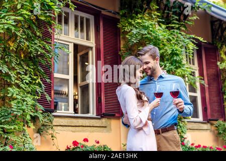 Glückliches junges Paar hält Gläser Rotwein beim Stehen in der Nähe von Haus bedeckt mit grünem Efeu Stockfoto