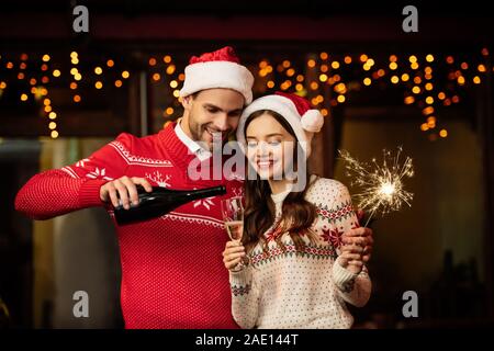 Fröhlicher Mann gießen Champagner im Glas von Happy Freundin holding Sparkler Stockfoto