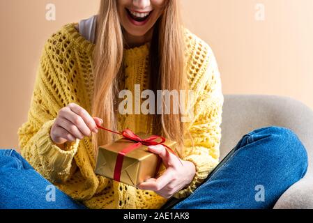 7/8-Ansicht von aufgeregt Frau in gelb Pullover holding Geschenkbox im Sessel sitzt auf beige Stockfoto
