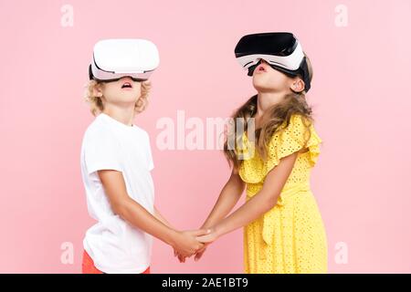 Kinder mit Virtual reality Headset halten sich an den Händen und sah auf rosa isoliert schockiert Stockfoto