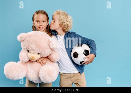 Junge mit Fußball küssen Freund mit Teddybär auf Blau isoliert Stockfoto