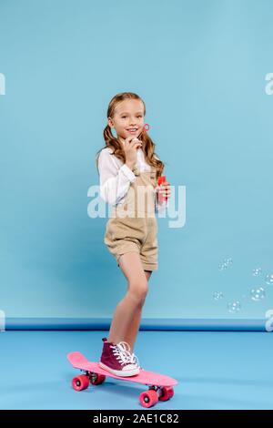 Lächelnd und cute kid stehend auf Penny board und bläst Seifenblasen auf blauem Hintergrund Stockfoto