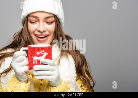 Happy girl in gelb Pullover, Schal, Mütze und Handschuhe holding Tasse Kakao isoliert auf Grau Stockfoto