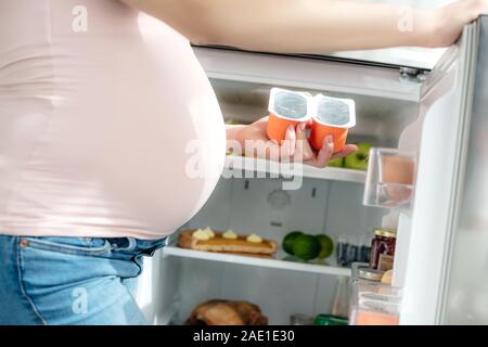 7/8-Ansicht der schwangeren Frau mit Joghurt in der Nähe von geöffneten Kühlschrank in der Küche Stockfoto