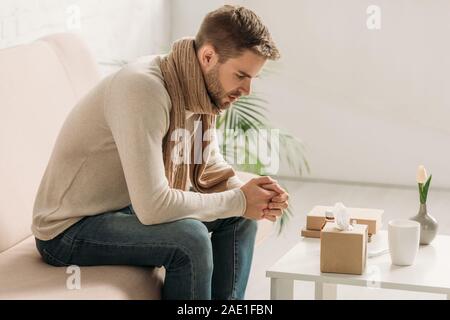 Kranken Mann in warmen Schal und Pullover sitzen auf einem Sofa in der Nähe von Tabelle mit Arzneimitteln Stockfoto