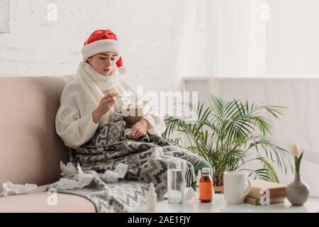 Kranke Frau in Santa Hut, in der Decke eingewickelt, sitzt auf einem Sofa in der Nähe von Tabelle mit Medikamenten und Thermometer Stockfoto