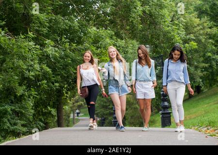 Vier attraktive junge Freunde Frauen sprechen und gemeinsam zu Fuß auf Gehweg mit Bäumen Stockfoto