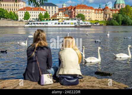 Zwei kaukasischen Frauen sitzen auf einem Felsen beobachten weiße Schwäne und Enten und Mähren Fähre der Moldau in Prag in der Tschechischen Republik Kreuzfahrt. Stockfoto