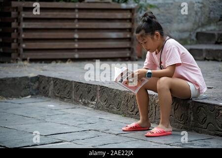 Feng Huang, China - August 2019: junge Chinesin auf einem Gehsteig und lesen Buch der Kinder sitzen, Feng Huang Altstadt Stockfoto