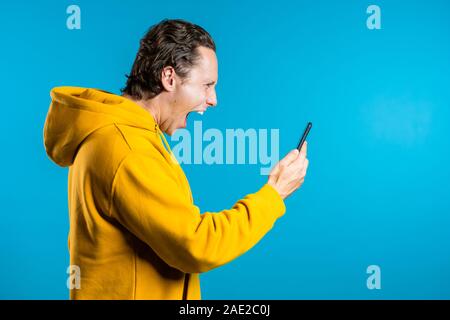 Zornigen Mann in gelb Hoodie, Schreien sein Handy. Gestresst und deprimiert Kerl schwört, mit jemandem auf blauem Hintergrund. Stockfoto