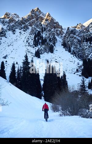 Radfahrer in Red Jacket Fahrten auf Schnee Straße in den Bergen bei Sonnenaufgang im Winter Stockfoto