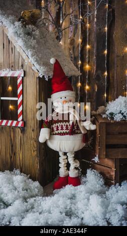 Ein großer Schneemann mit rotem Hut und weißen Schal, der in ein elegantes rotes Licht, steht in der Nähe ein Holzhaus auf einem Winter Weihnachten Hintergrund. Einrichtung in einem Foto st Stockfoto