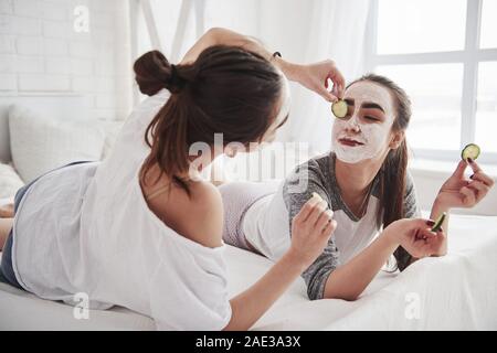 Das wird großes auf sie schauen. Konzeption der Hautpflege mit weißen Maske und Gurken auf dem Gesicht. Zwei weibliche Schwestern haben Wochenende im Schlafzimmer Stockfoto