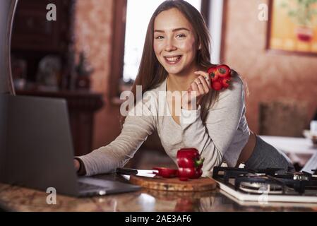 Die Scheibe der roten Pfeffer und lächelnd. Hübsche junge Frau, die in der modernen Küche in der Nähe von Gas- Herd und mit Laptop Stockfoto