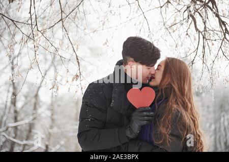 Halten roten herzförmigen Pappe. Wunderschöne junge Paare haben gute Zeit zusammen im verschneiten Wald Stockfoto
