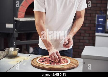 Professional auf seine Arbeit. Baker in weißem Hemd, Oliven lecker Pizza für eine Bestellung im Restaurant zu machen Stockfoto