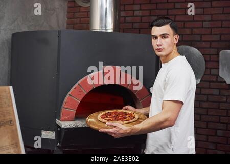 Stilvolle Baker in Weiß tragen, zieht die Pizza aus dem Ofen Stockfoto