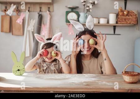 Halten Sie Eier in der Nähe der Augen. Mutter und Tochter im bunny Ohren zu Ostern haben Sie etwas Spaß in der Küche tagsüber Stockfoto