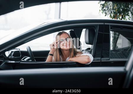 Grüne Pflanze hinter sich. Schöne blonde Mädchen in der neuen Auto sitzen mit modernen schwarzen Interieur Stockfoto