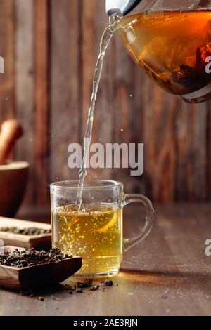 Frischer grüner Tee wird aus einer Teekanne, die in einem Becher gegossen. Hot grüner Tee im Glas Tasse auf einem Holztisch. Stockfoto