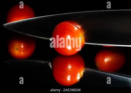 Trauben Tomaten vor einem schwarzen Hintergrund mit einem scharfen Messer auf einer reflektierenden Oberfläche in Scheiben geschnitten wird. Lebendige Farben und Raum für Kopie Stockfoto