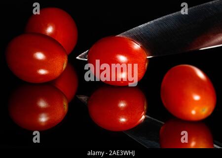 Trauben Tomaten vor einem schwarzen Hintergrund mit einem scharfen Messer auf einer reflektierenden Oberfläche in Scheiben geschnitten wird. Lebendige Farben und Raum für Kopie Stockfoto