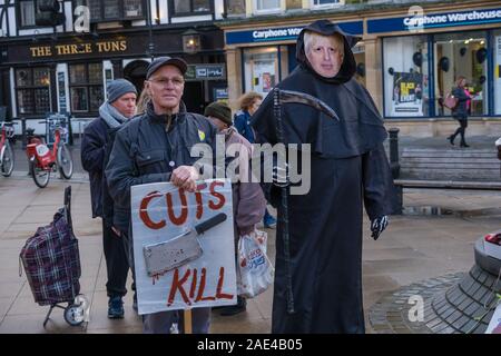 London, Großbritannien. 6. Dezember 2019. Ein Mann mit einem BorisJohnson Maske verkleidet als der Sensenmann steht neben anderen ein Plakat mit einem Cleaver und die Meldung 'Schnitte an der Straße Theater trial Töten' durch behinderte Menschen gegen Sozialabbau (DPAC) im Zentrum von Uxbridge Aufruf auf die Wähler Boris Johnson und die Tories abzulehnen. Sie verweisen auf die geschätzten 200.000 vorzeitige Todesfälle durch Kürzungen und Sanktionen einschließlich der Beseitigung der Independent Living Fund, zu Universal Kredit, zu einem enormen Anstieg der Foodbank verwenden, Hunger, Verschuldung und Zwangsräumungen und ein UN-Bericht von "schwerwiegenden und systematischen Menschenrechtsverletzungen geführt hat, Stockfoto