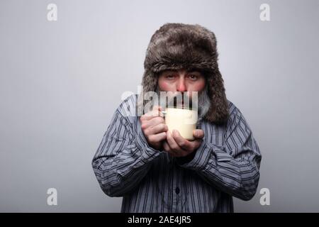 Kranken Mann mit Schlafanzug und mit einer Kappe weht eine heiße Tasse Tee auf einem grauen Hintergrund Stockfoto