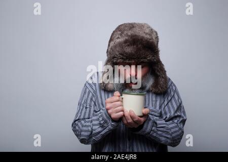Grippe kranken Mann mit Schlafanzug und mit einer Kappe weht eine heiße Tasse Tee auf einem grauen Hintergrund. Gesundheit Konzept. Stockfoto