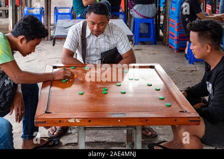 Zwei burmesische Teens spielen das Brettspiel Carrom auf einem Holzbrett mit einem anderen jungen Mann beobachtet das Spiel in einem Markt, in Mandalay, Myanmar (Birma) Stockfoto