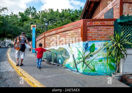 Vater und Sohn gehen durch ein Wandbild einer großen grünen Leguan auf einem Backsteingebäude in Puerto Vallarta. Der junge weist auf die Iguana. Stockfoto