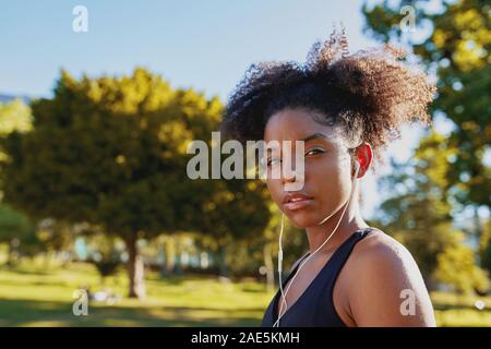 Porträt eines Fitness afrikanische amerikanische junge Frau mit Kopfhörern in den Ohren zu Musik im Park zu hören, bevor sie Übung an einem sonnigen Tag Stockfoto