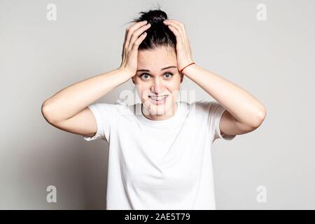 Durch eine unerwartete Überraschung schockiert, die Frau lächelt seltsam und unsicher, und sieht in die Kamera, ihre Hände auf den Kopf. Gemischte Gefühle und Stockfoto