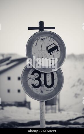 Zwei alte schmutzige Verbotszeichen Verkehrsschild: überholverbot oder vorbei und die Geschwindigkeitsbegrenzung. Haus und Snowy Mountains in Abend im Hintergrund. Schwarz und Weiß Stockfoto