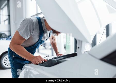 Unter der Haube. Mann in der blauen Uniform und dem schwarzen Hut Reparatur beschädigter Automobil Stockfoto