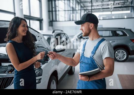 Achten Sie beim nächsten Mal. Frau im Auto Salon mit Mitarbeiter in der blauen Uniform, Ihrem reparierten Auto zurück Stockfoto