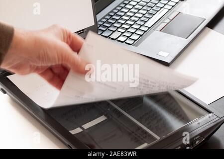 Eine Hand mit einem Stück Papier auf einen Flachbettscanner mit einem Laptop im Hintergrund. Selektiver Fokus und Hand in Bewegungsunschärfe. Stockfoto