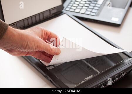 Eine Hand mit einem Stück Papier auf einen Flachbettscanner mit einem Laptop im Hintergrund. Selektiver Fokus Bild. Stockfoto