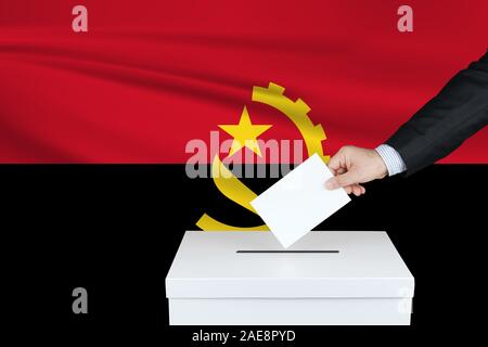 Wahl in Angola. Die Hand des Menschen, der seine Stimme in die Wahlurne setzt. Angola Flagge auf Hintergrund gewellt. Stockfoto